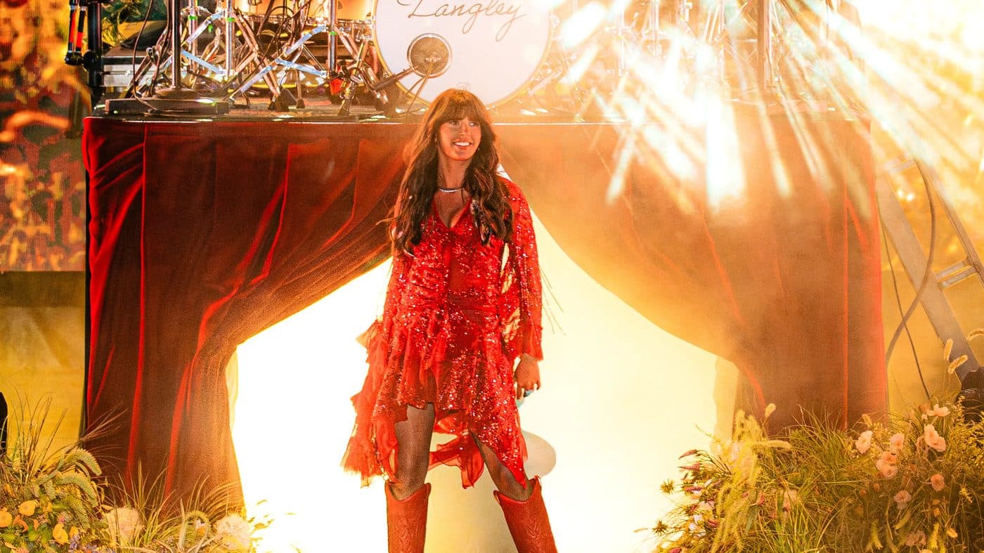 Ella Langley walking out through a red stage curtain in a red fringed dress and boots under a burst of golden light on the Dandelion Tour
