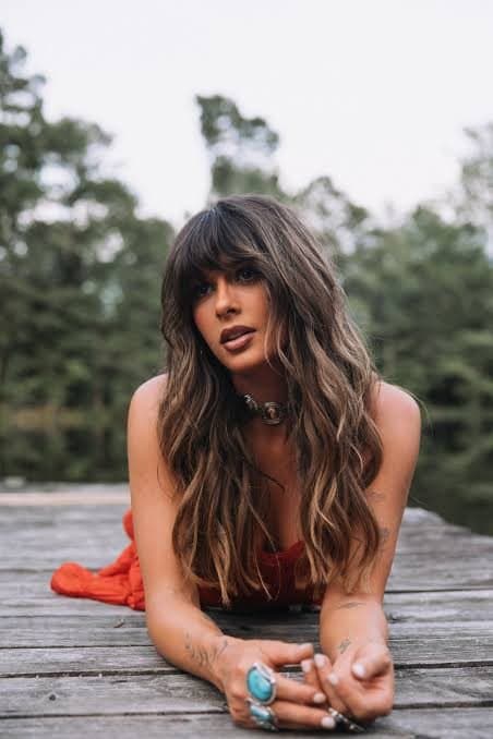 Ella Langley lying on a wooden deck outdoors in a red top, propped on her elbows and looking into the camera