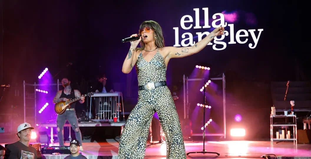 Ella Langley singing with one arm raised in a leopard-print jumpsuit and sunglasses, her name lit on the backdrop behind the band