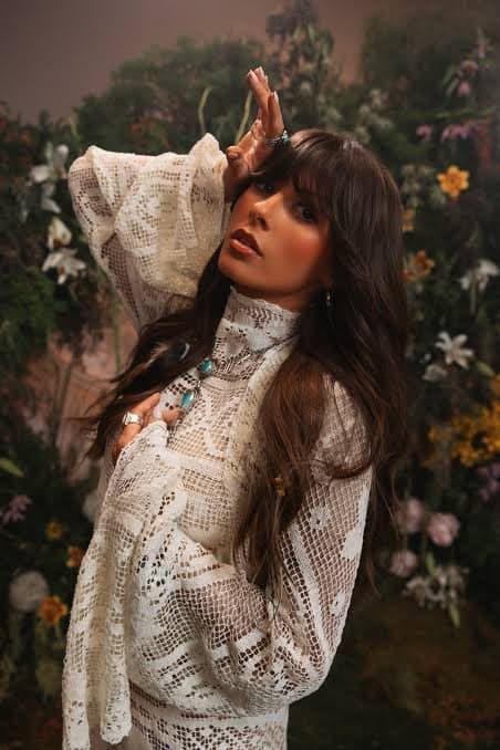 Ella Langley in a white lace dress with one hand raised to her hair, posed against a wall of flowers