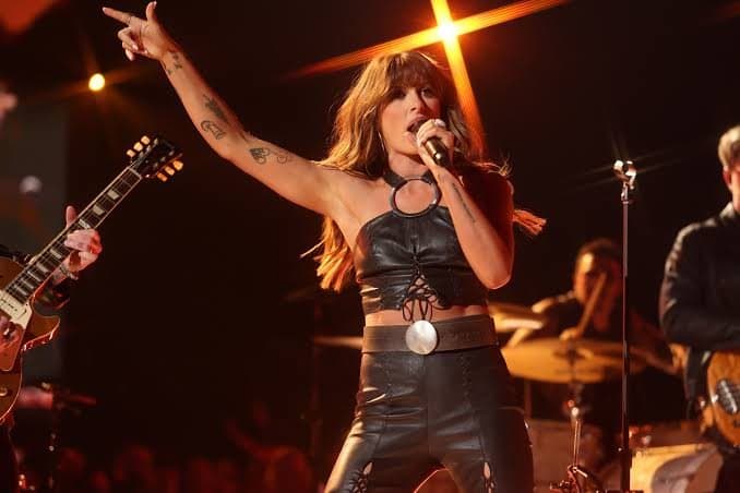 Ella Langley in a black leather outfit and concho belt with one arm raised to the crowd, guitarist behind her in warm light